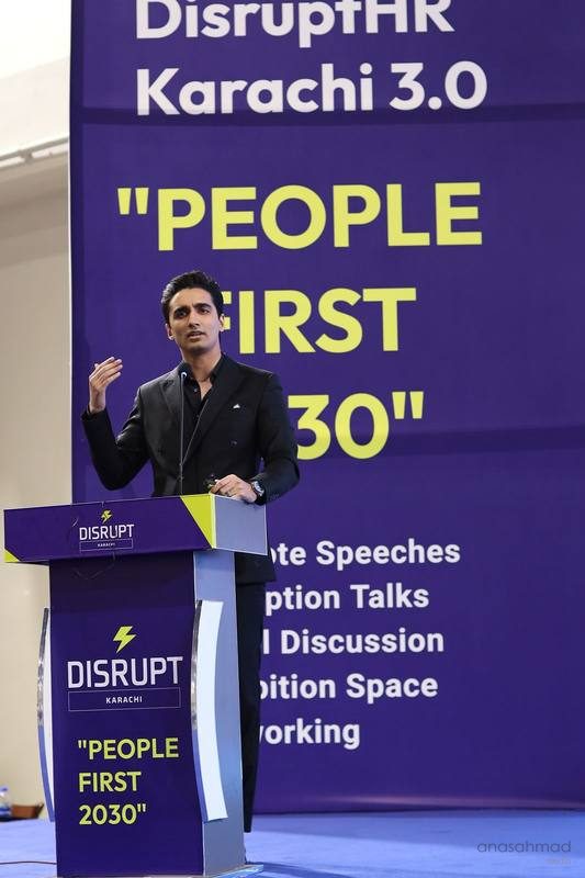 Man speaking at a podium on stage at a DisruptHR Karachi conference