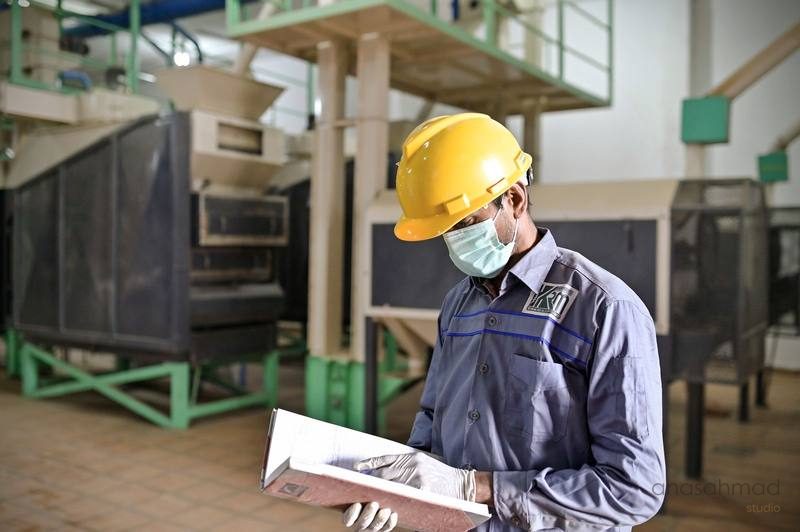 Factory worker in hard hat and mask inspecting a textile production line with a clipboard — Anas Ahmad Studio, Karachi