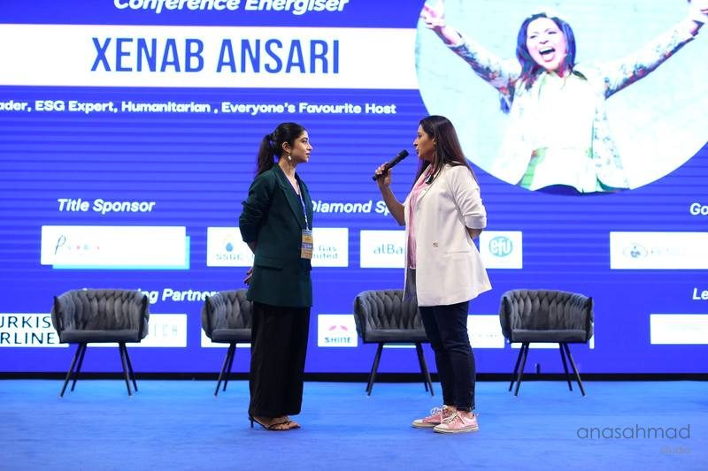 Conference host interviewing a speaker on stage in front of a large screen and sponsor logos