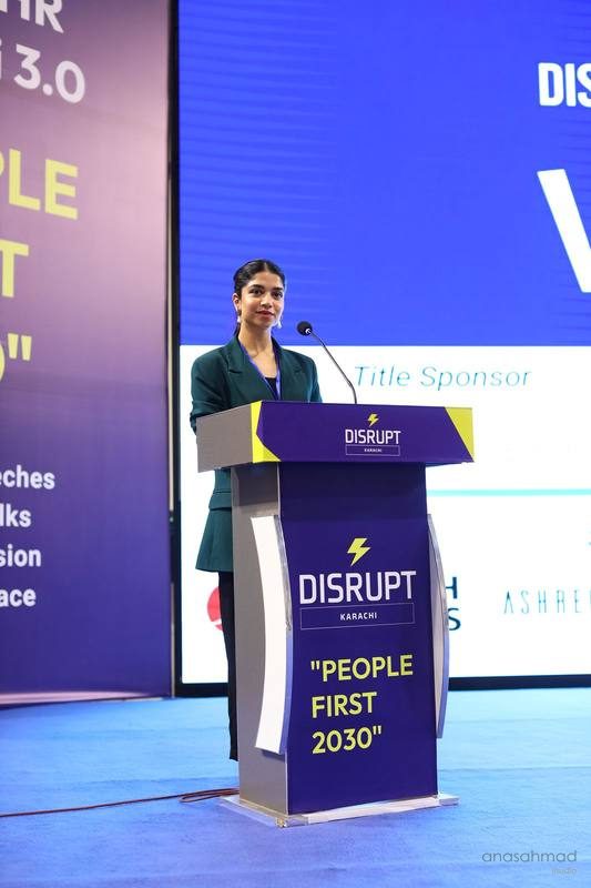 Woman speaking at a podium on stage at a DisruptHR Karachi conference