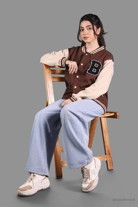 Female model in brown and cream varsity jacket with wide-leg jeans, seated on wooden chair